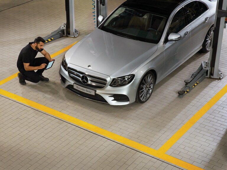 Un técnico de Mercedes-Benz se encuentra arrodillado frente a un vehículo, mientras realiza la revisión principal.